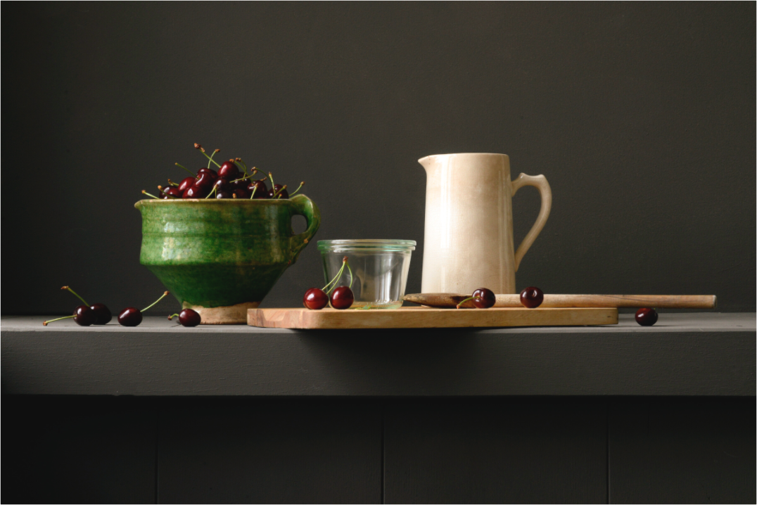 Stilleven met groene kom vol kersen, roomkleurige kan, glazen pot en houten lepel op donkere achtergrond.