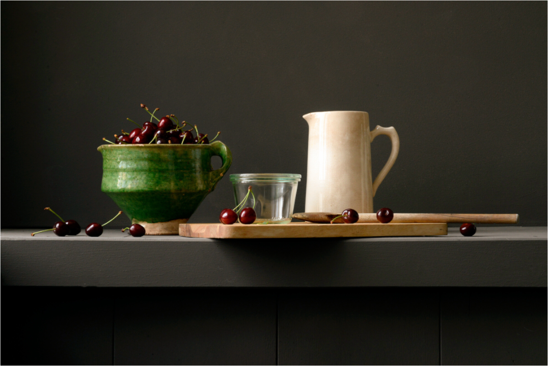 Stilleven met groene kom vol kersen, roomkleurige kan, glazen pot en houten lepel op donkere achtergrond.