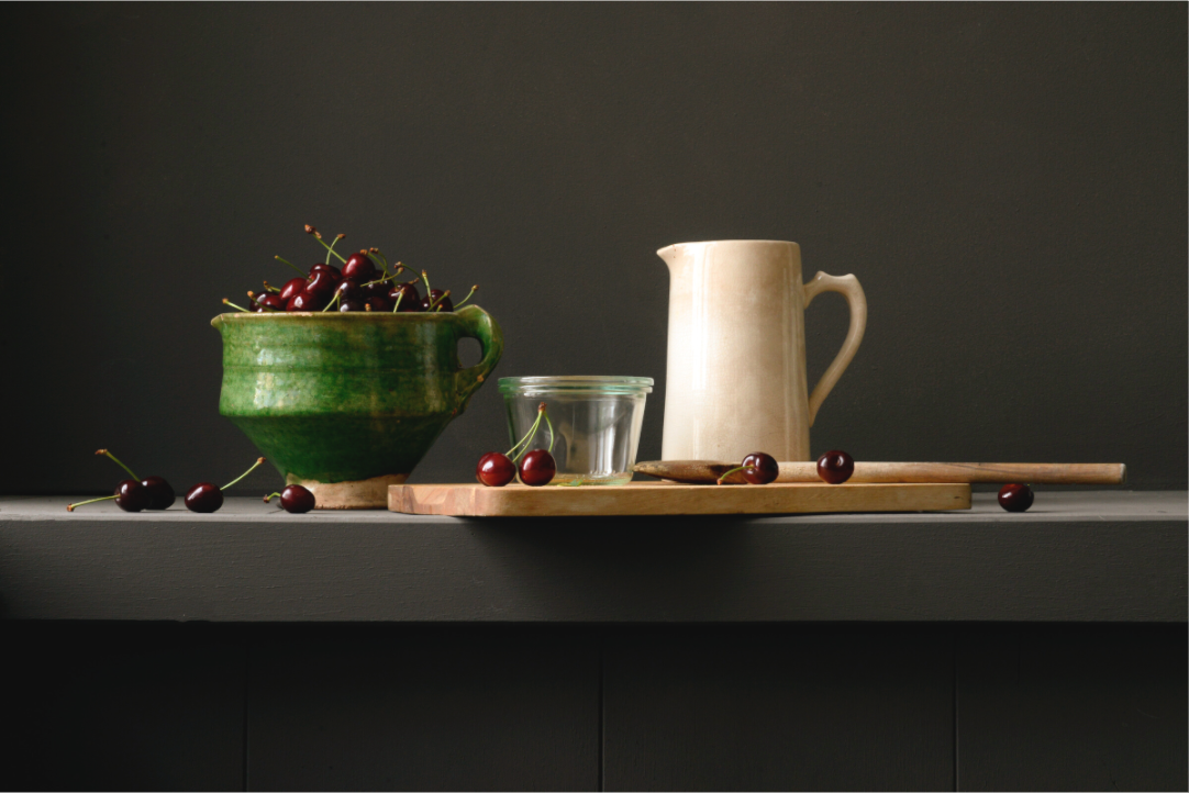Stilleven met groene kom vol kersen, roomkleurige kan, glazen pot en houten lepel op donkere achtergrond.