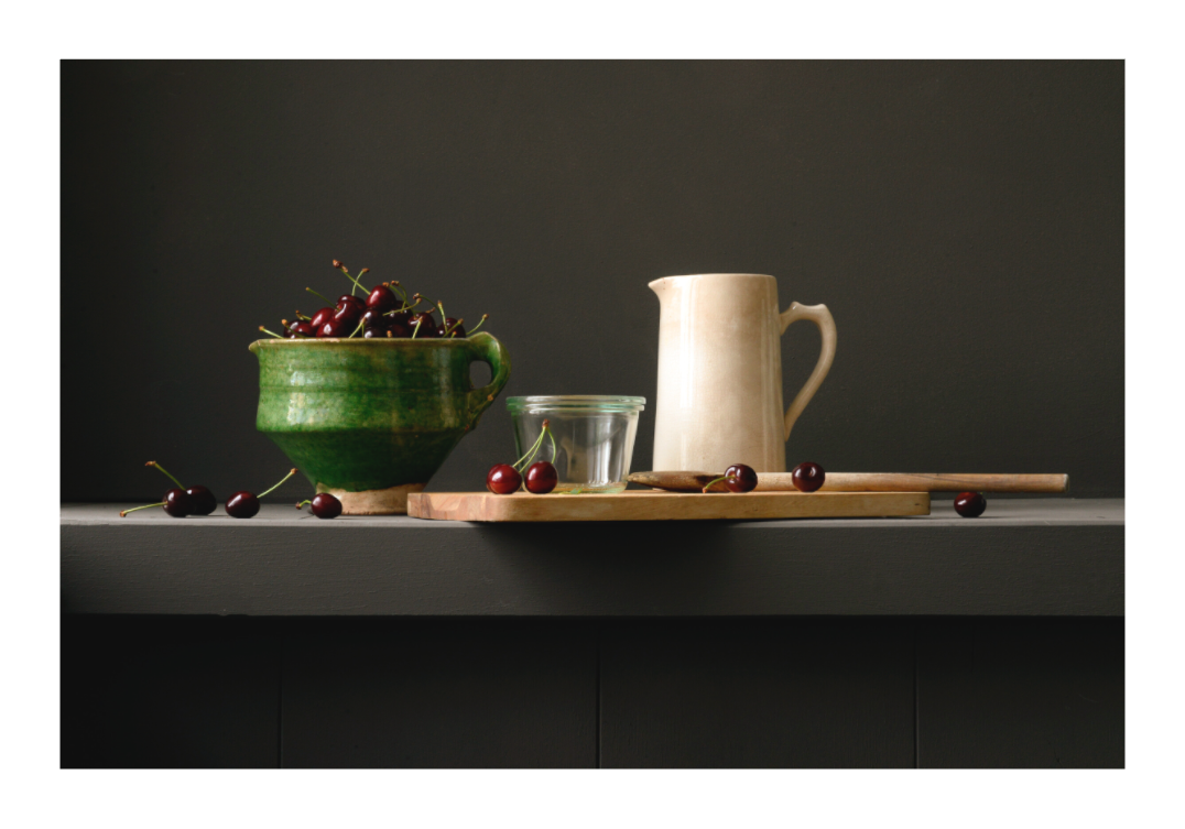 Stilleven met groene kom vol kersen, roomkleurige kan, glazen pot en houten lepel op donkere achtergrond.