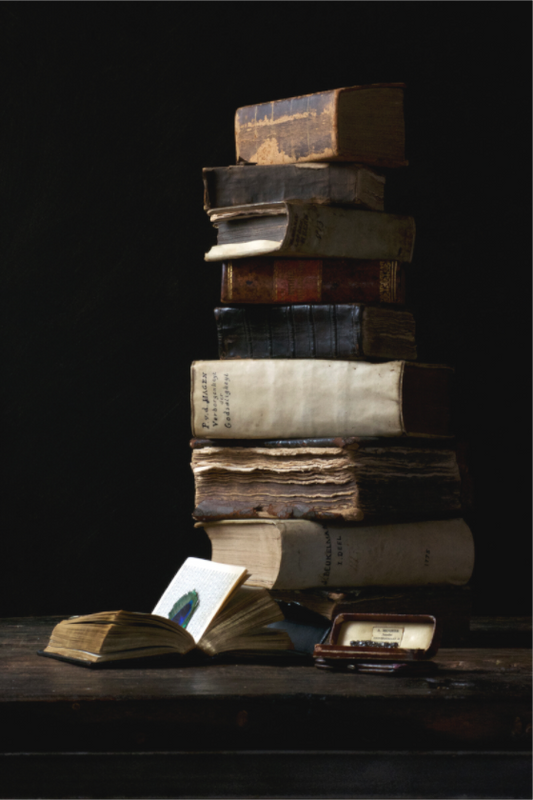 Stilleven van een hoge stapel oude boeken, open boek met pauwenveer en klein kistje op een donker houten blad. Fotografie van Dirk Bal.