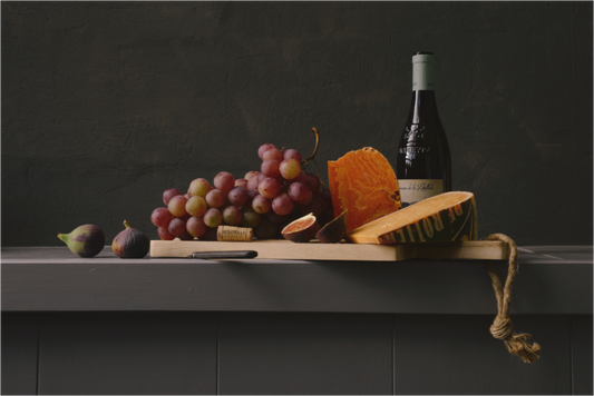 Realistisch stilleven met rode druiven, kaas, vijgen en wijnfles op houten plank, gefotografeerd door kunstenaar Dirk Bal tegen een donkere achtergrond.